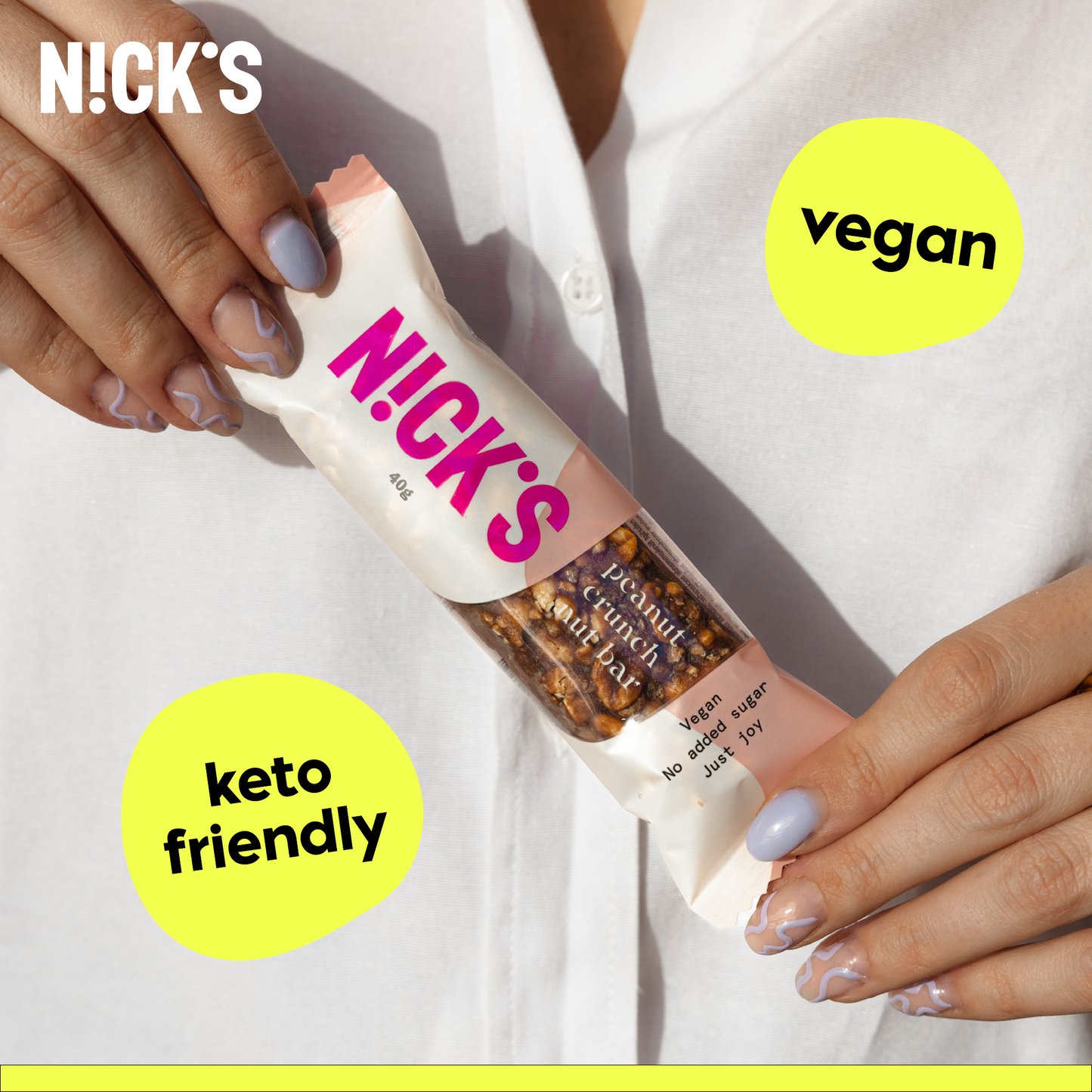 Close-up of hands with painted nails holding a N!CK'S Peanut Crunch Nut Bar. Text highlights 'keto friendly' and 'vegan' features of the snack.